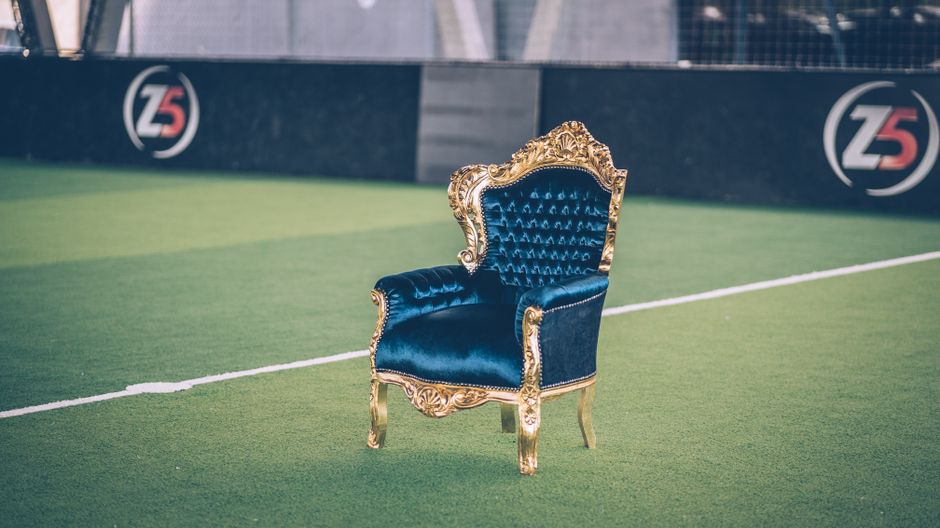 An elegant antique armchair with gold ornate wooden frame and dark blue velvet upholstery, positioned on a green sports field with artificial turf. The chair features tufted backrest and seat, with in