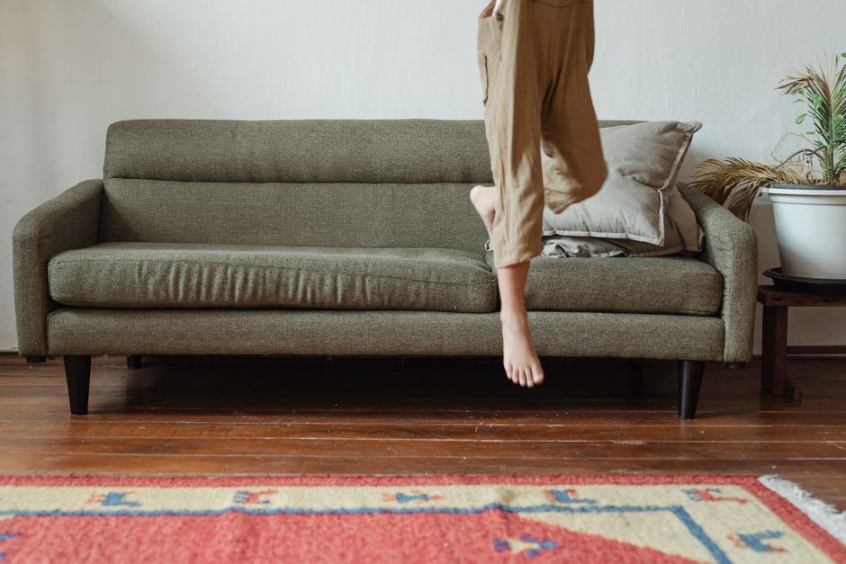 A beige fabric sofa with tufted cushions and a wooden base, located against a plain white wall in a domestic living room. The sofa appears to be in clean condition, with no visible stains or dirt, und